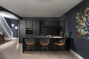 Una cocina moderna con encimera oscura, tres sillas de estructura metálica y un cuadro abstracto grande en la pared.