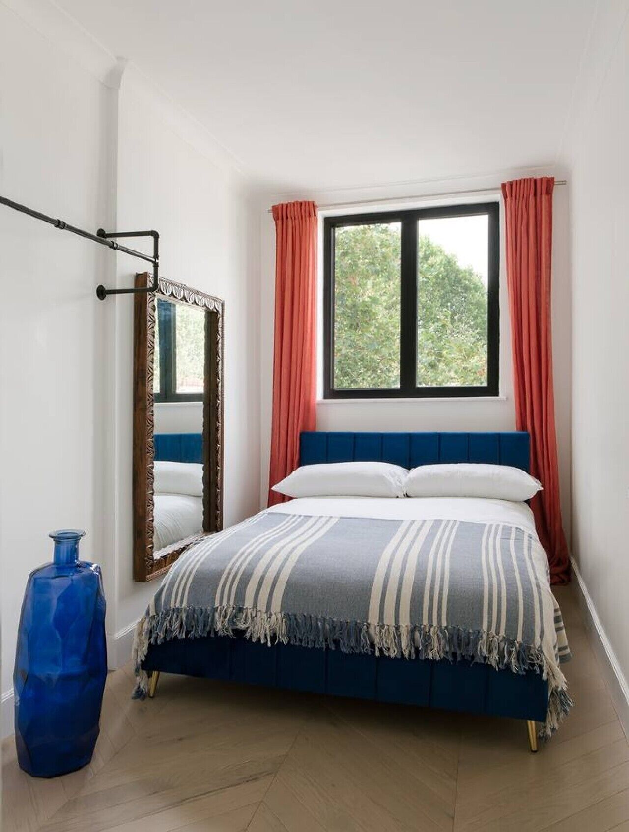Un dormitorio con una cama, una ventana con cortinas, un espejo y un jarrón azul.