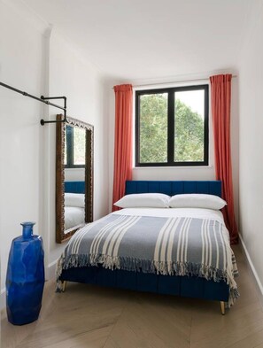 Ein Schlafzimmer mit einem Bett, einem Fenster mit Vorhängen, einem Spiegel und einer blauen Vase.