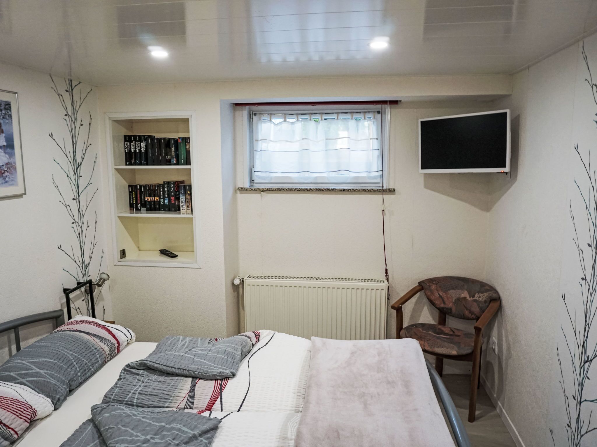 Un dormitorio con una cama, una estantería, un radiador, una ventana con cortinas, una silla y un televisor fijado a la pared.