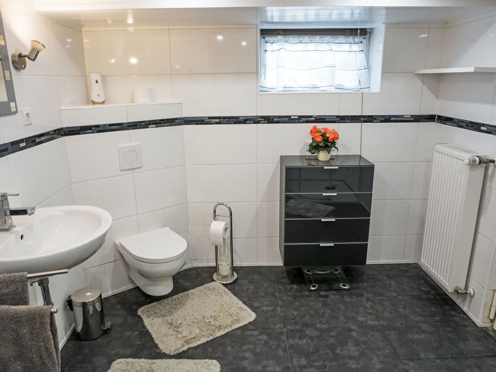 A modern bathroom with a white sink, toilet, and black storage unit, a small window, and a radiator.