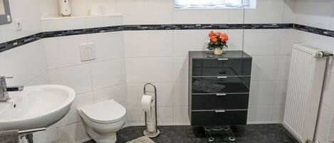 A modern bathroom with a white sink, toilet, and black storage unit, a small window, and a radiator.