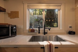 A kitchen with a stainless steel sink, a microwave, a window with a view of trees, and a wooden wall map.