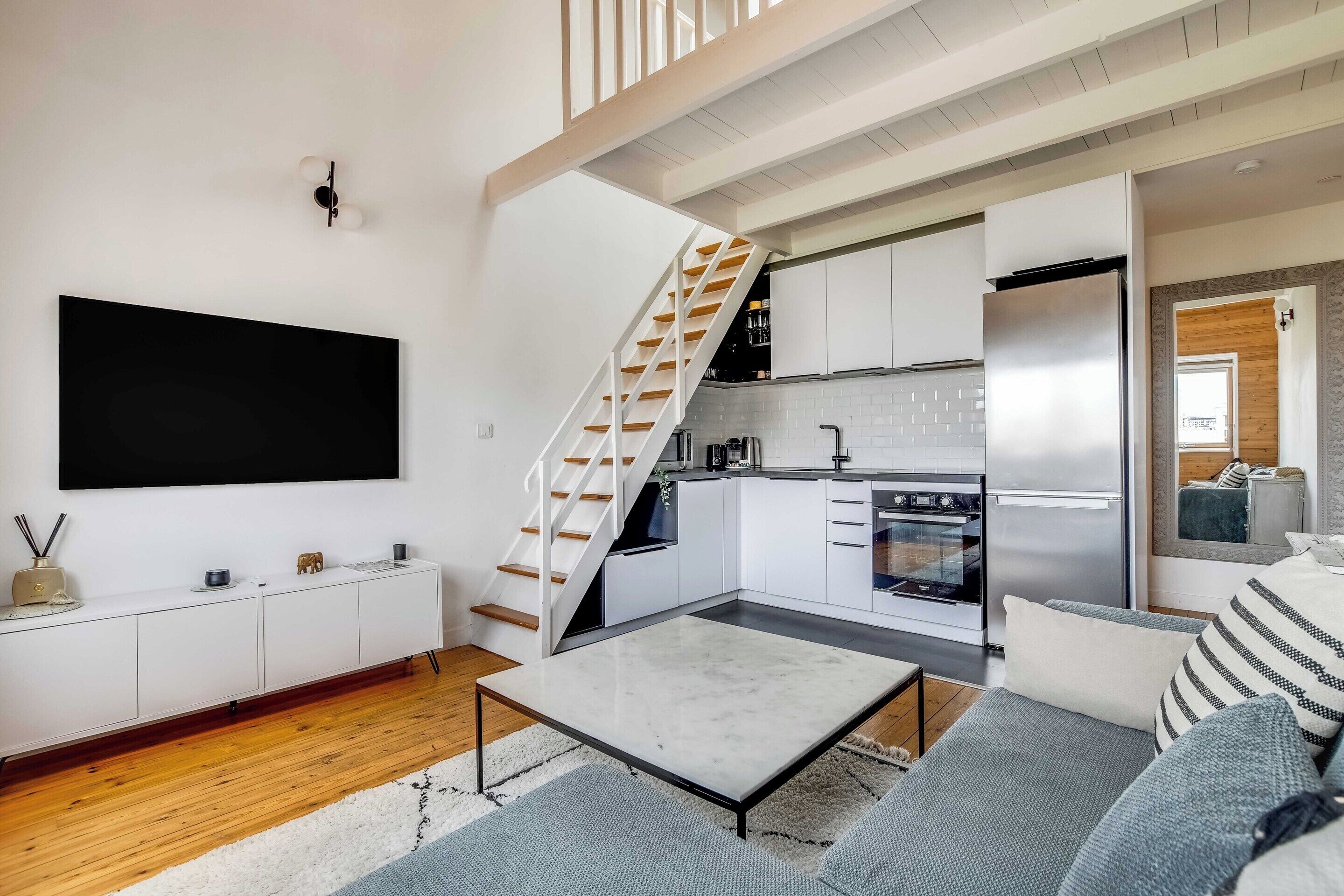 A modern living room with a flat-screen TV, a sofa, a coffee table, and a staircase leading to a kitchen.