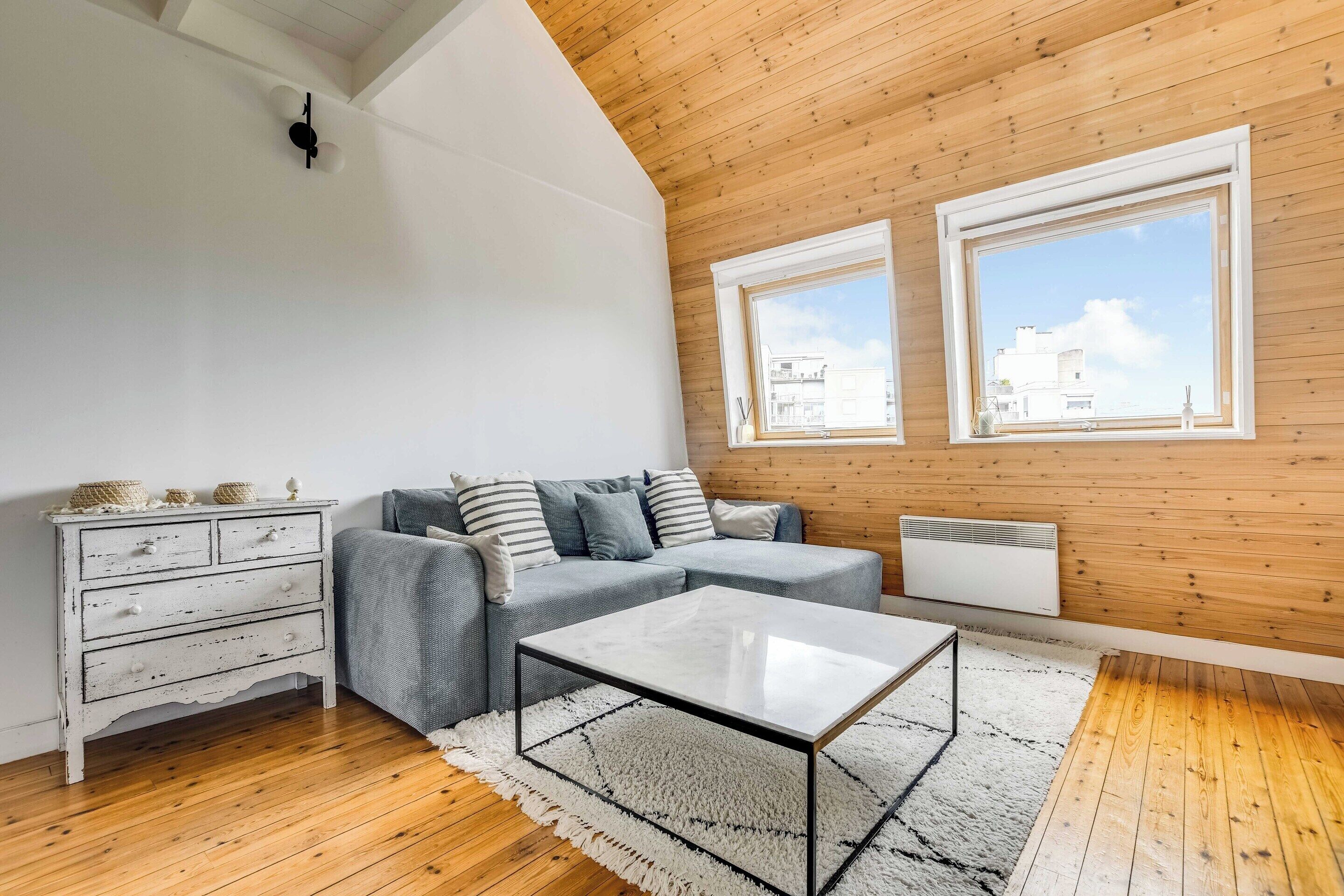A loft-style room with a wooden ceiling, a grey sofa, a white dresser, a glass coffee table, and a view of buildings through two windows.
