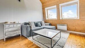 Ein Loft-Zimmer mit Holzbalkendecke, einem grauen Sofa, einer weißen Kommode, einem Glastisch und Blick auf Gebäude durch zwei Fenster.