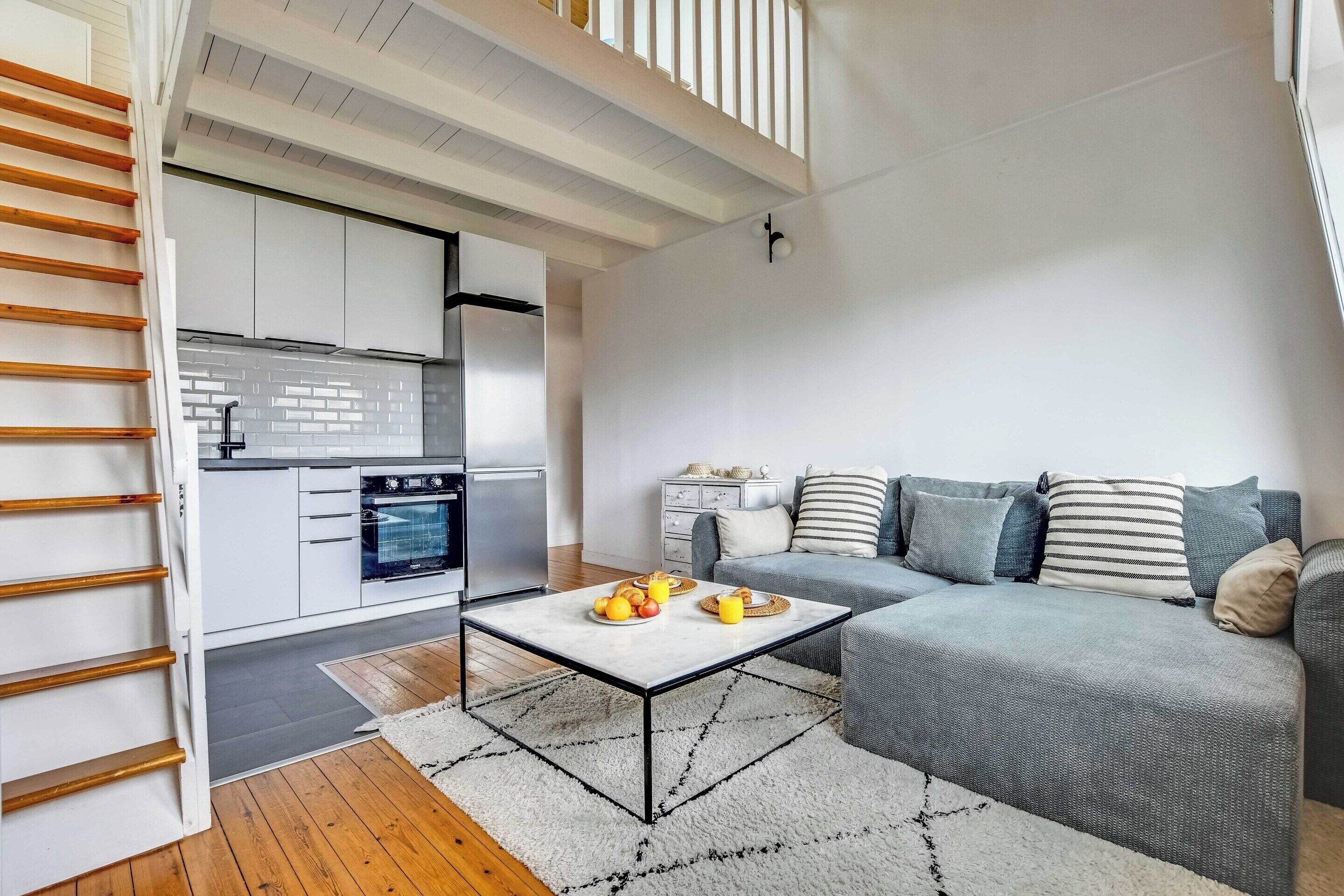 A modern living room with a grey sofa, a coffee table with fruits, a kitchen with white cabinets, and a wooden staircase.