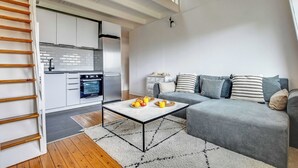 Ein modernes Wohnzimmer mit einer grauen Couch, einem Couchtisch mit Früchten, einer Küche mit weißen Schränken und einer Holztreppe.