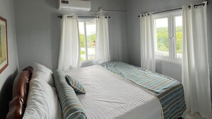 Een slaapkamer met een houten plafond, een bed met wit bedlinnen, twee ramen met gordijnen, en een airconditioning.