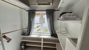 Een kleine, lichte kamer met een raam, een bankje en een plank met opgevouwen kleding.
