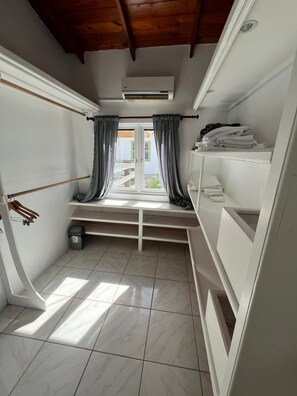 Ein kleiner, heller Raum mit Fenster, einer Bank und einem Regal mit gefalteter Kleidung.