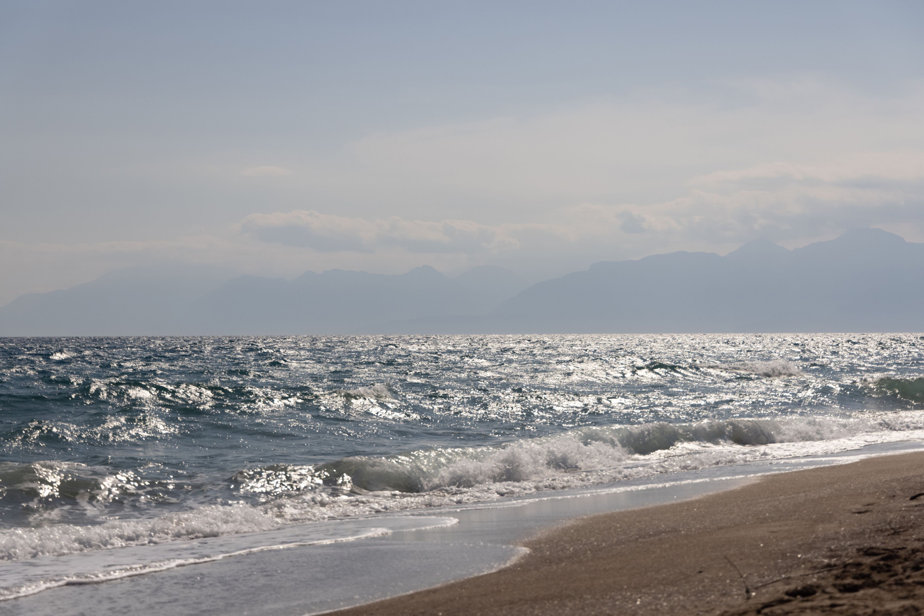 Une plage avec des vagues, un ciel dégagé et des montagnes à l’horizon.