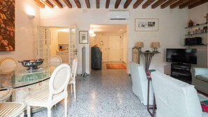 Un'ampia zona giorno con tavolo da pranzo, sedie bianche, divano, TV e cucina sullo sfondo.