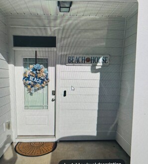 Eine Tür mit einem Schild, das 'BEACH HOUSE' zeigt, eine Fußmatte und ein Fenster mit einem Kranz.