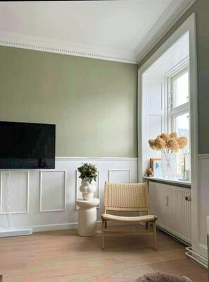 Ein Zimmer mit Fernseher, einem Korbstuhl, einer weißen Vase mit Blumen und einem Fenster mit Blick.