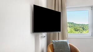 Une pièce avec un téléviseur fixé au mur, une fenêtre donnant sur un paysage verdoyant et un fauteuil marron avec un coussin.