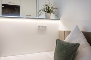 A bedroom with a bed, a bedside table, a nightstand, a wall socket, and a potted plant.