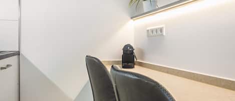 A modern room with a white wall, a black chair, a small table, a potted plant, and a wall-mounted light fixture.