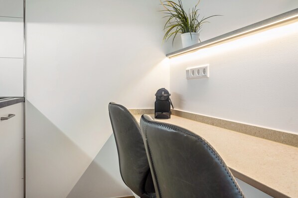 A modern room with a white wall, a black chair, a small table, a potted plant, and a wall-mounted light fixture.