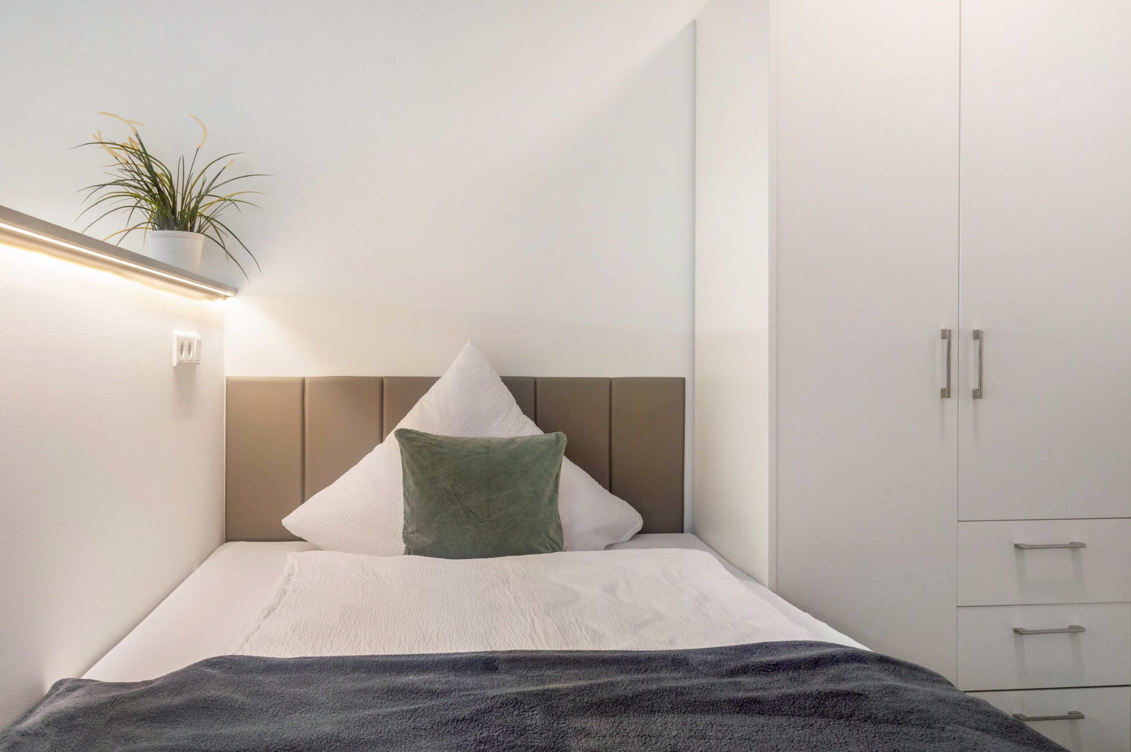 Quarto minimalista com uma cama, um travesseiro verde, um travesseiro branco, uma planta na mesa de cabeceira e um guarda-roupa.