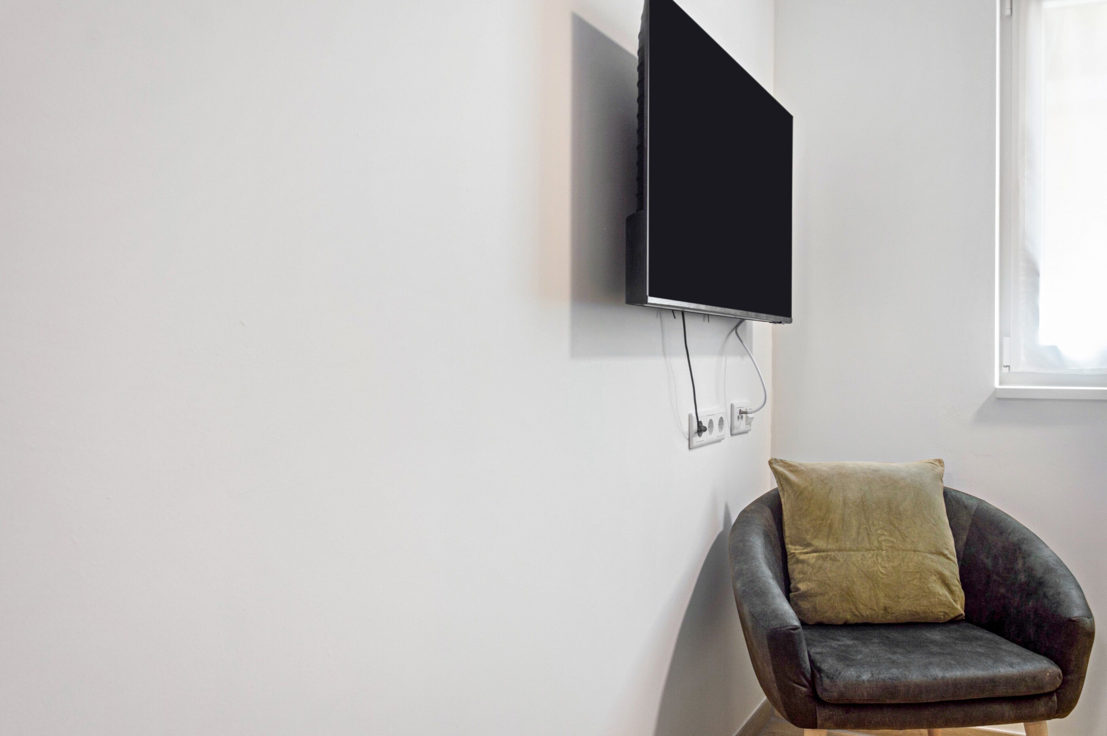A modern living room with a mounted flat-screen TV, a single armchair with a cushion, and a window providing natural light.