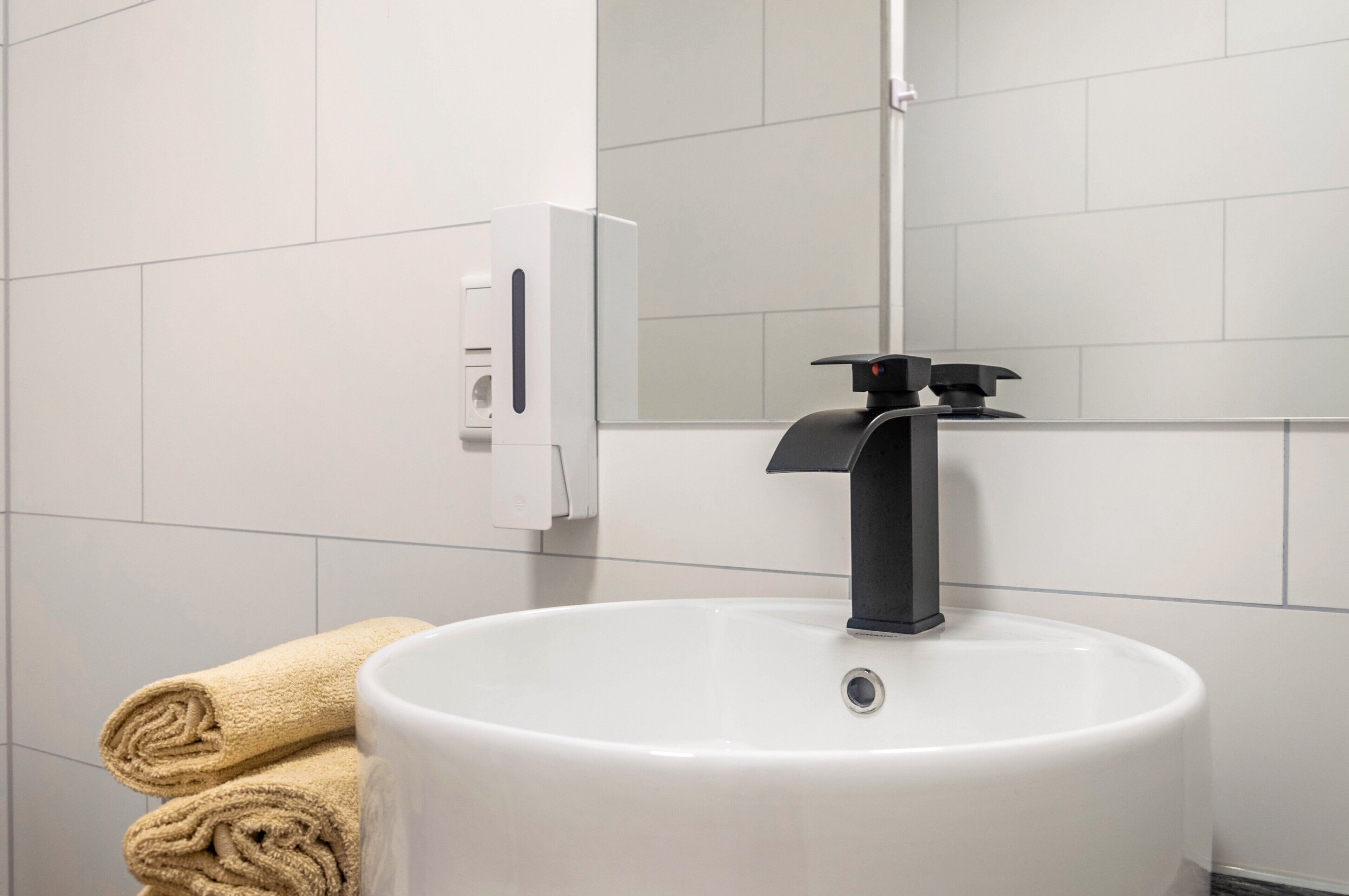 A bathroom with a white sink, a black faucet, a towel dispenser, and a mirror.