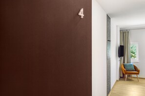 Une pièce avec un mur marron, une enseigne portant le chiffre « 4 », un plancher en bois, une chaise avec un coussin et une fenêtre avec des rideaux.