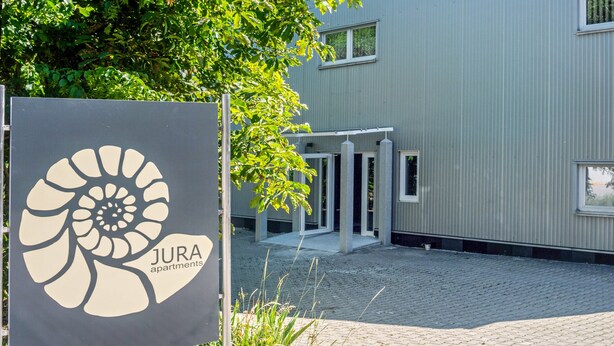 Skilt for JURA lejligheder med en moderne bygning i baggrunden.