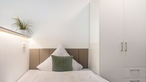 A neatly made bed with a headboard, a green pillow, and a white pillow. There is a plant on a shelf above the bed, and a white wardrobe with drawers to the right. - Studio Apartment '7' with Wi-Fi (Parsberg)