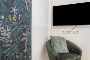 Ein Zimmer mit einem an der Wand befestigten Flachbildfernseher, einem grünen Sessel mit Kissen und einer Wand mit Blättermuster.