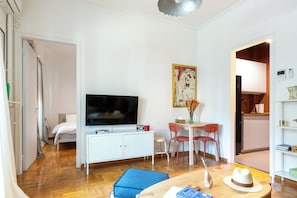 Ein kompakter Wohnbereich mit Bett, Fernseher, Essecke mit Stühlen, einem kleinen Tisch und einer Küchenzeile.