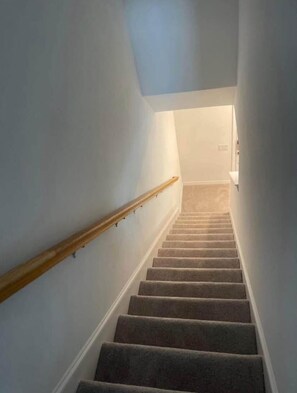A carpeted staircase with a wooden handrail, white walls, and a light fixture at the top.