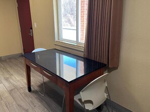 A room with a glass-top table, a white chair, a wooden door, and a window with curtains. - Bragg Motel (Upper Marlboro)