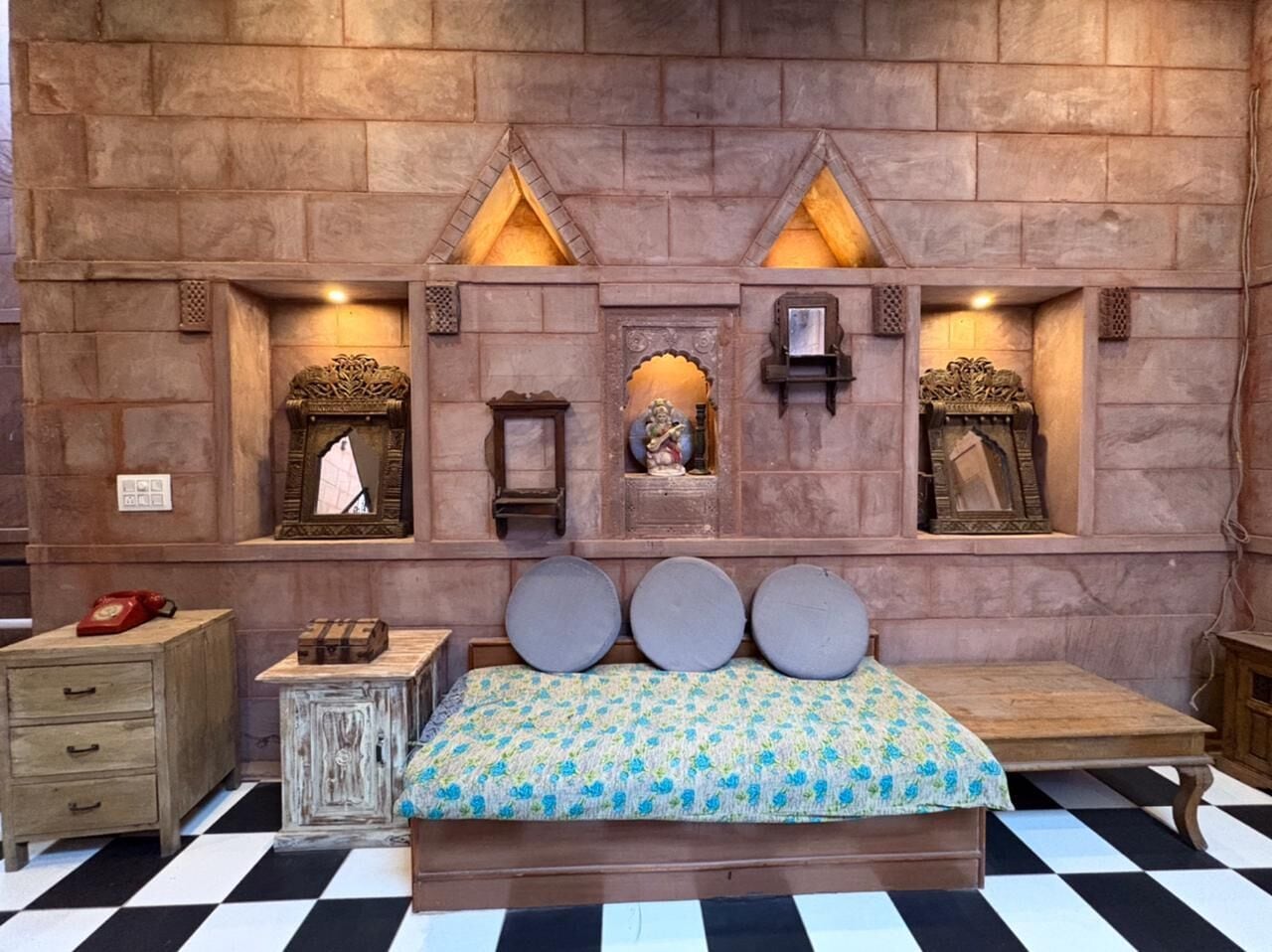 Una habitación con piso a cuadros, una cama con colcha estampada, una mesita de noche de madera, un aparador y una pared con estantes empotrados que contienen objetos decorativos.