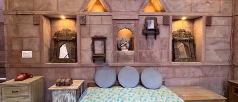 A room with a checkered floor, a bed with a patterned bedspread, a wooden nightstand, a dresser, and a wall with built-in shelves containing decorative items.