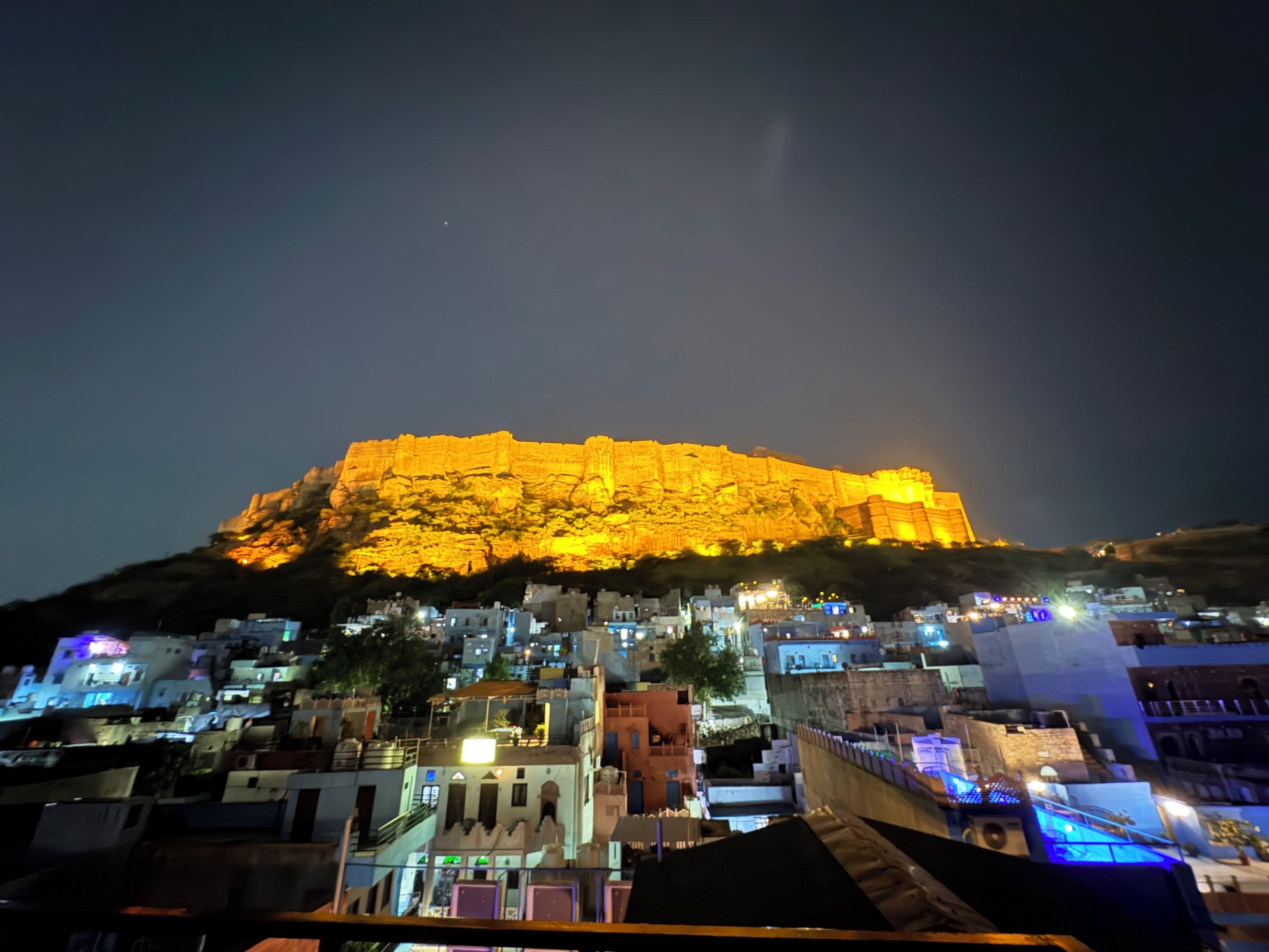 Un fort historique illuminé la nuit, entouré d’une ville densément bâtie où l’on aperçoit les toits et les bâtiments.