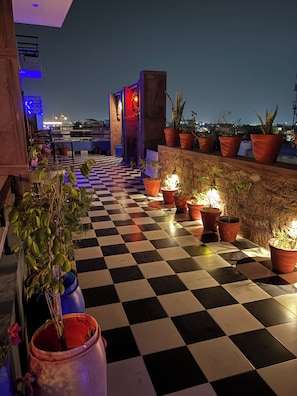 Una terraza en la azotea con suelo a cuadros, plantas en macetas y vista panorámica de la ciudad de noche.