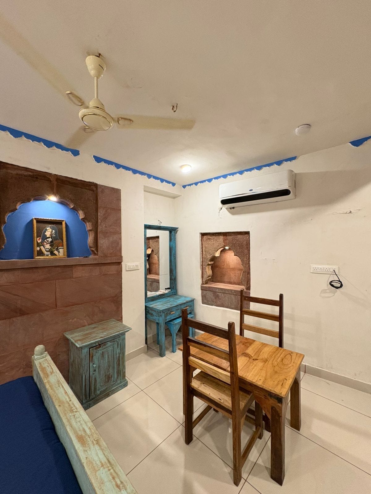 Una habitación con ventilador de techo, espejo con marco azul, silla de madera, una mesita pequeña y aire acondicionado instalado en la pared.