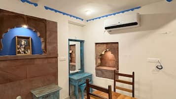 Una habitación con ventilador de techo, espejo con marco azul, silla de madera, una mesita pequeña y aire acondicionado instalado en la pared.