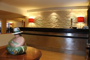 Un área de recepción con pared de piedra, mostrador de recepción, dos lámparas rojas y una estatua decorativa sobre una mesa.