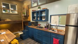 Een compacte keuken met blauwe kastjes, een houten aanrechtblad, een roestvrijstalen koelkast en een raam met jaloezieën.