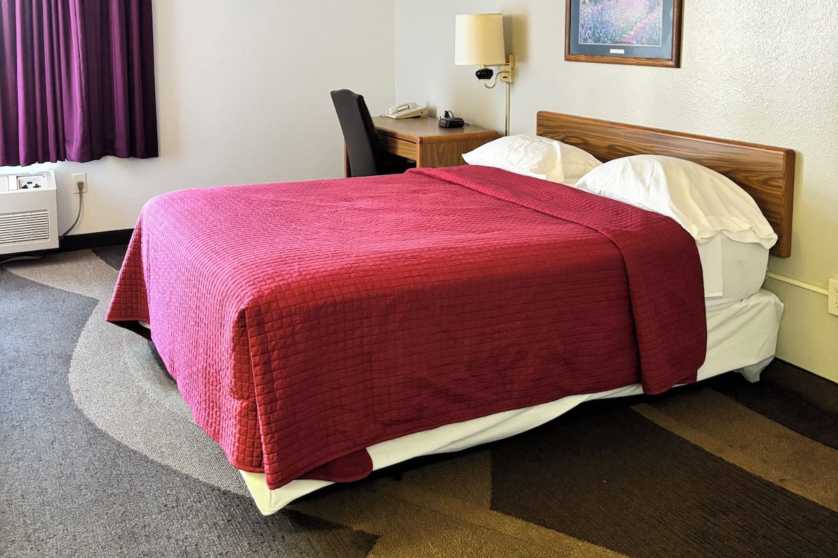 A hotel room with a bed, a desk, a chair, a lamp, and a painting on the wall.