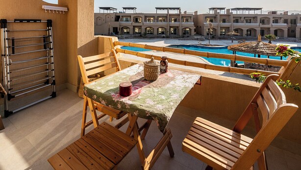 Een houten zitgedeelte buiten met een tafel en stoelen, een zwembad en een gebouwencomplex op de achtergrond.