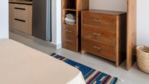 Een slaapkamer met een bed, een houten garderobe, en een zichtbare keuken door een open deur, plus een geweven mand.
