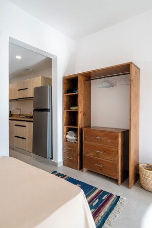 Ein Schlafzimmer mit Bett, einem Holzschrank, einer durchgehenden Tür erkennbaren Küche und einem gewebten Korb.