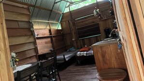 Interior rústico de una cabaña de madera con techo alto, una zona de comedor con sillas de madera, una cama y una cocina con fregadero y armarios.