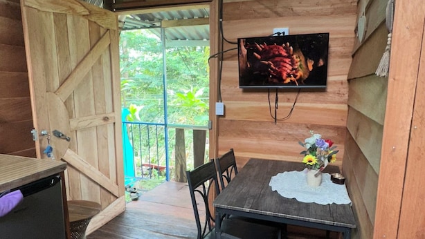 Interior de una cabaña de madera con una mesa de comedor, sillas, un televisor montado en la pared y una puerta que da al exterior.
