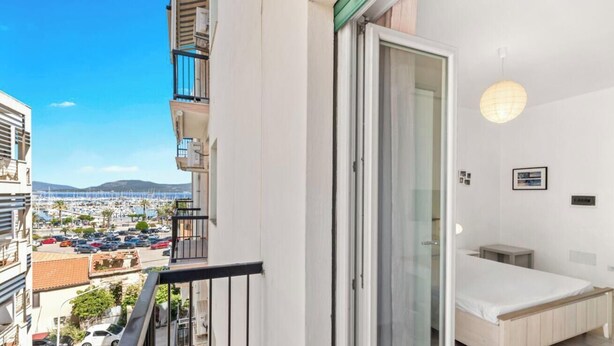 Ein Balkon mit Blick auf einen Hafen und Gebäude, eine Schiebetür, ein kleiner Nachttisch und eine Wandleuchte.