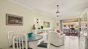 A dining area with a round table and chairs, a kitchen with white cabinets, and a view of a courtyard with a covered table and chairs. - ‘Férias Vilamoura’ Apartment with Balcony, Wi-Fi and Air Conditioning (Quarteira)