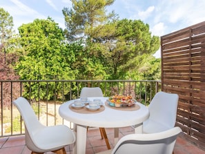 A round table with four white chairs, a small fruit bowl, and a plate of pastries on a balcony with a wooden fence and trees in the background. - Sant Eloi by Interhome (Tossa de Mar)